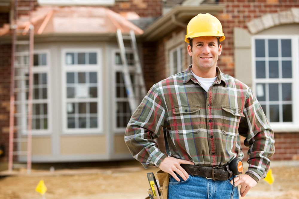 Construction worker in plaid shirt and hard hat, standing in front of a house under construction, smiling.