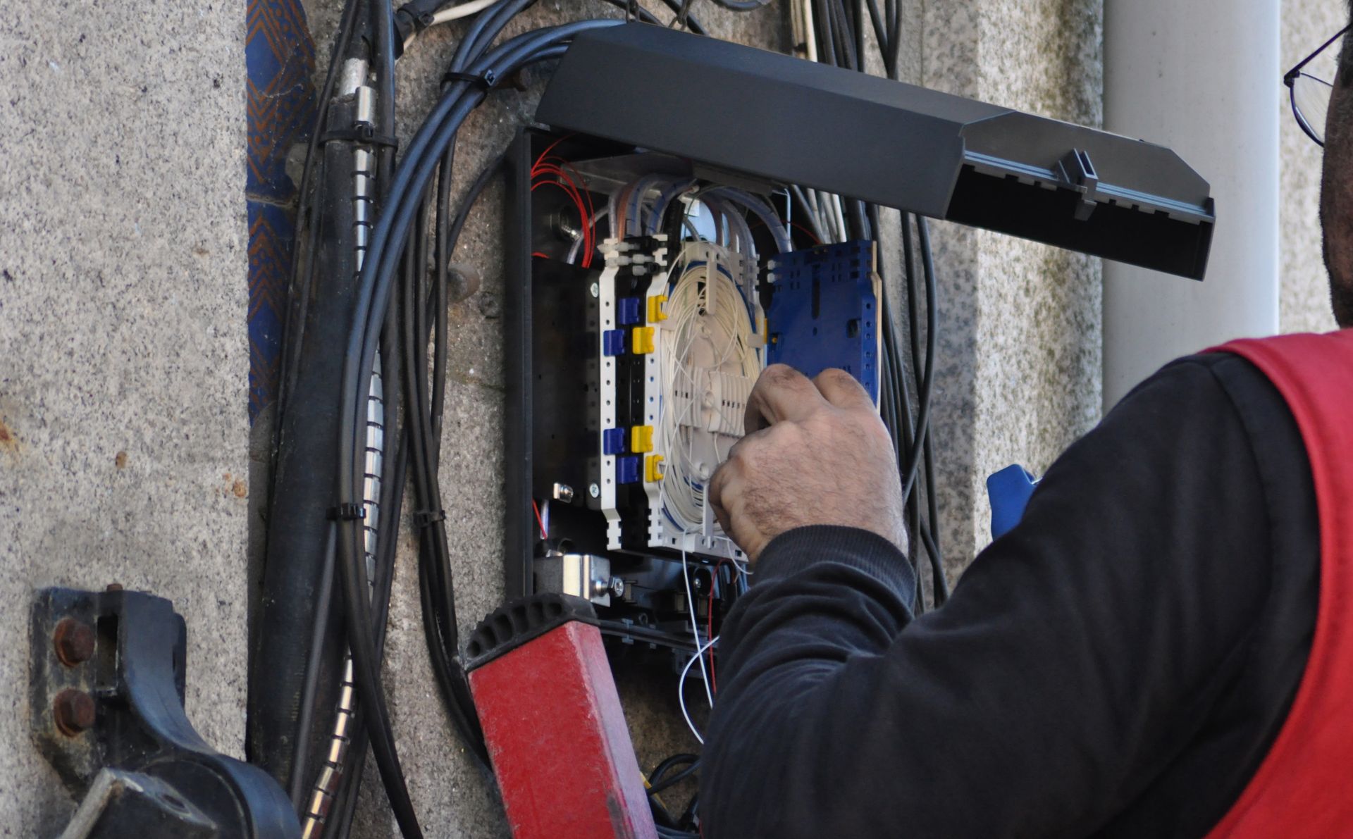 Person working on a network box mounted on a wall. Black cables, blue and yellow components visible.