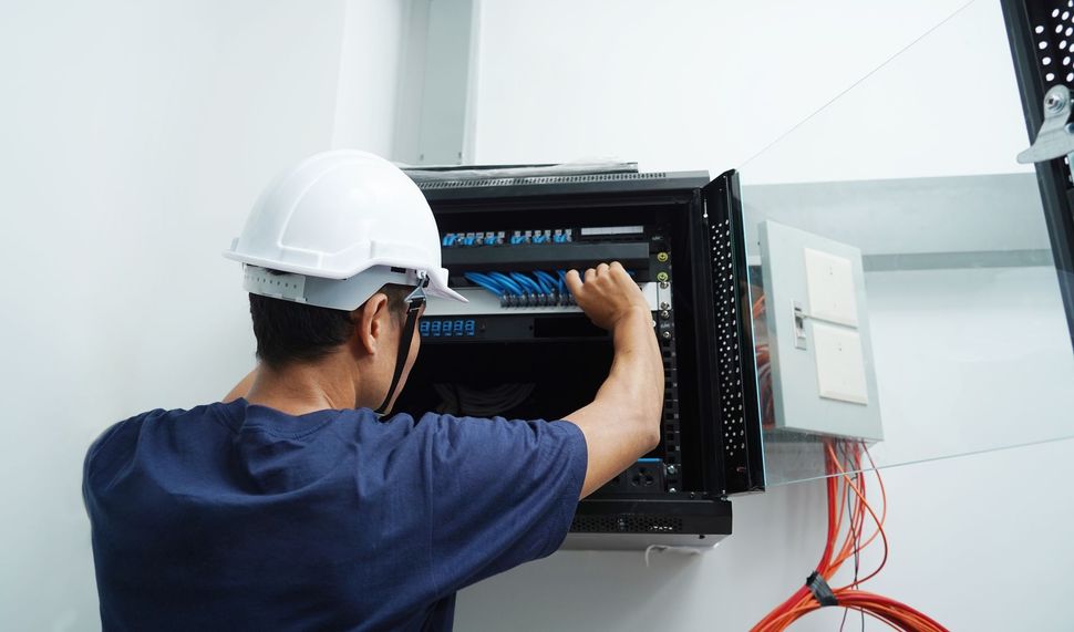 A technician wearing a white hard hat installs network cables into a wall-mounted server rack in a bright room.