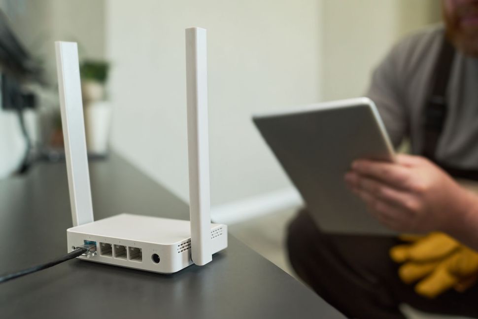White Wi-Fi router with antennas on a table, a person holding a tablet is in the background.