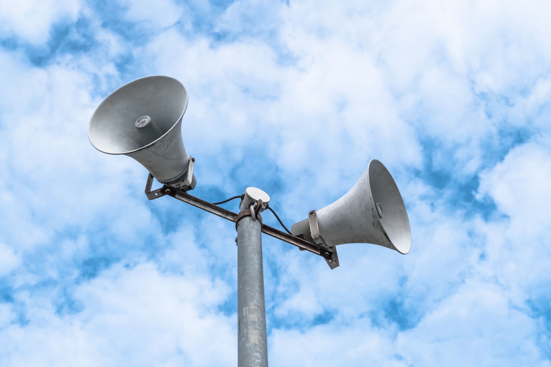 Two gray loudspeakers mounted on a pole against a blue, cloudy sky.