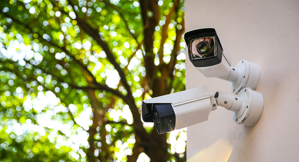 Two white security cameras mounted on a light-colored wall against a blurred background of green trees.