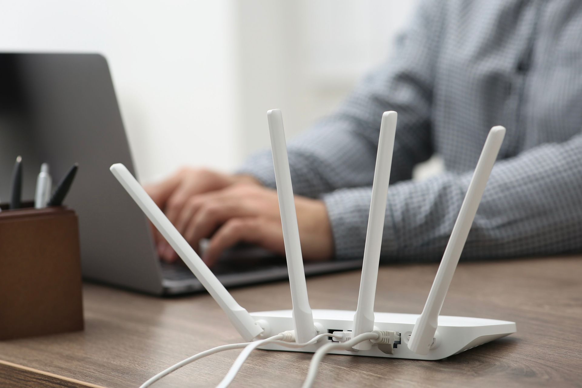 White router with four antennas in front of a person using a laptop on a wooden desk.