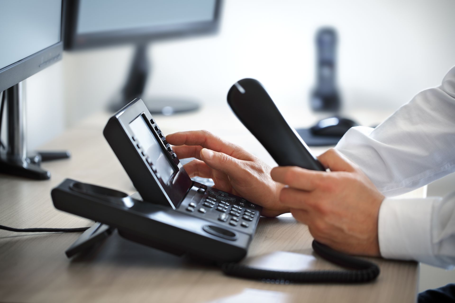 Hands holding a phone receiver near a desk phone with a display and number pad in an office setting.