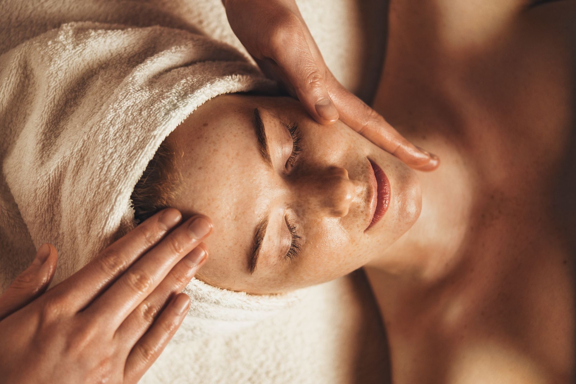 A woman with a towel wrapped around her head is getting a facial massage.