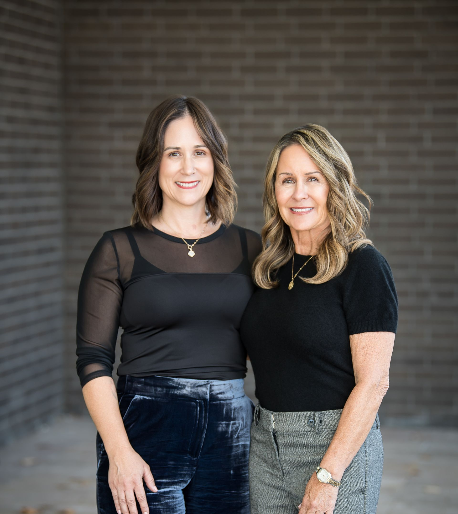 Two women standing in front of a brick wall, both wearing black tops and smiling.