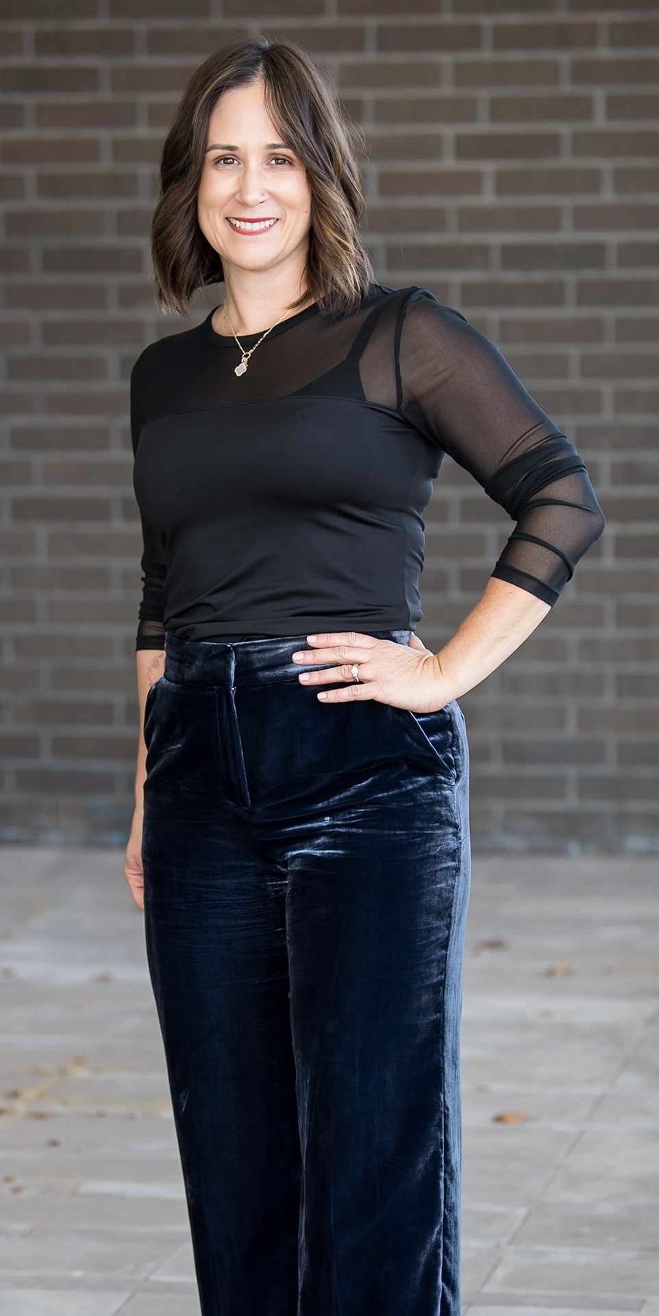 Two women posing together; one wears a black top and blue velvet pants, the other a black top and patterned gray pants.