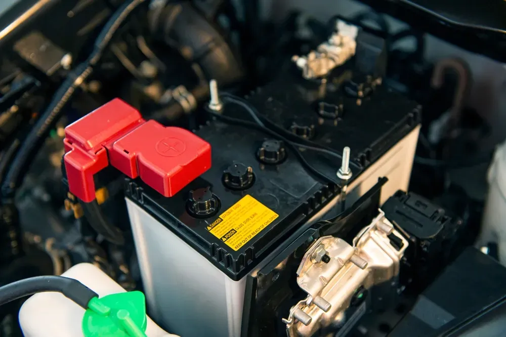A car battery sits inside an engine bay, with a red plastic cover over the positive terminal and a yellow warning label. — Murwillumbah Motor Wreckers in Byron Bay, NSW