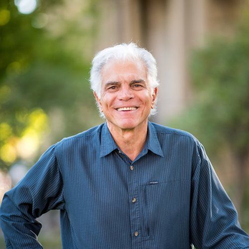 A man in a blue shirt is standing with his hands on his hips and smiling.