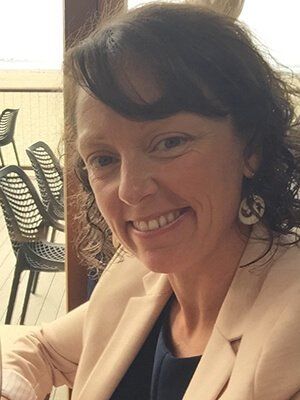 A woman is smiling while sitting at a table with chairs in the background.