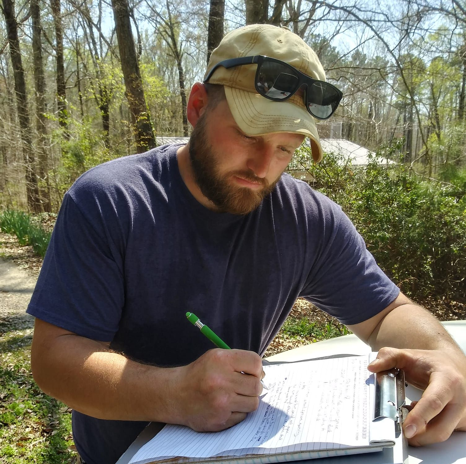 Stephen in cap and sunglasses writing on a clipboard outdoors in a wooded area