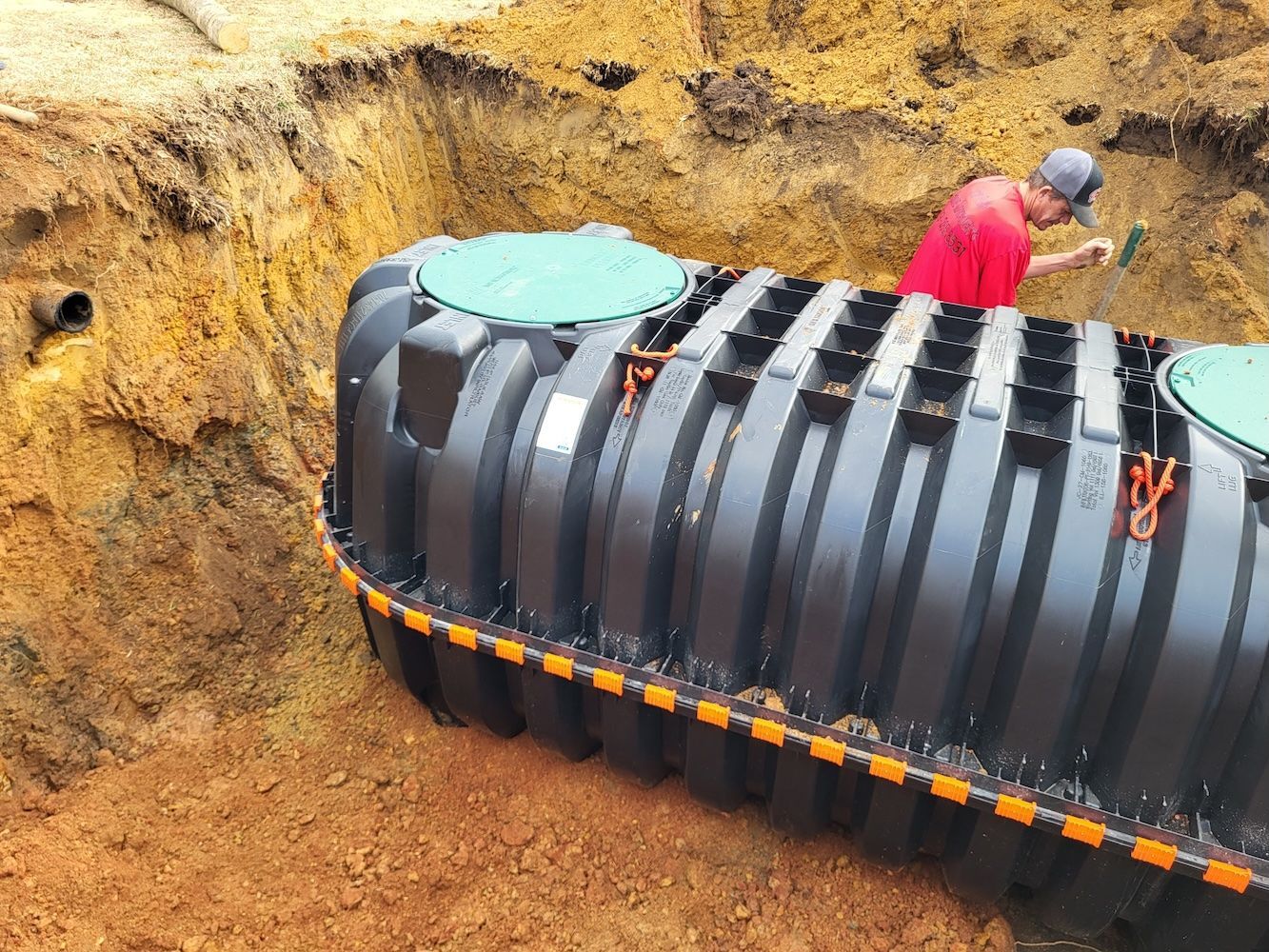 Underground black tank being installed in a dug-out dirt pit by a worker in a red shirt