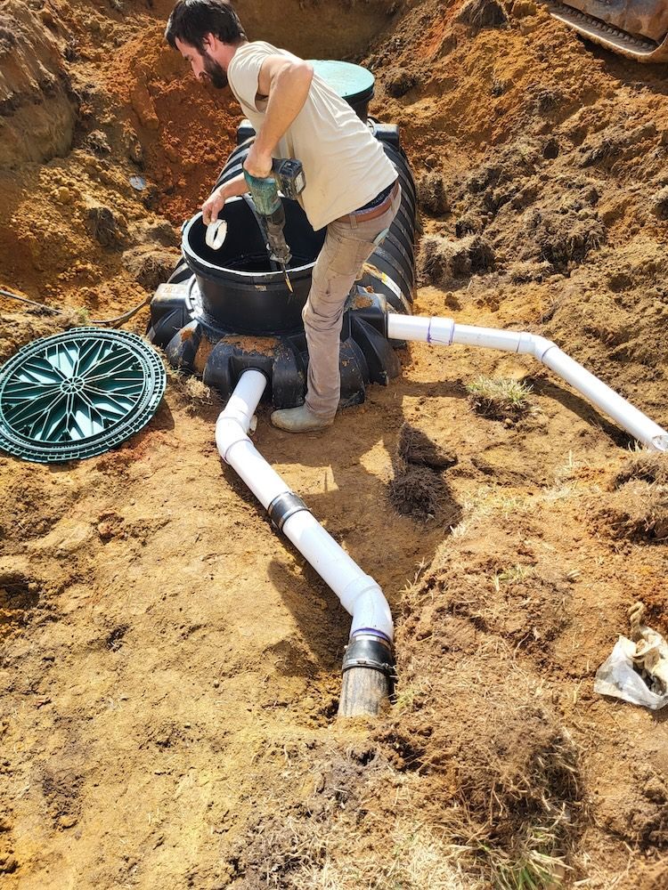 Worker installing black plastic pipe in an excavation pit with white drainage pipes and a green cover nearby