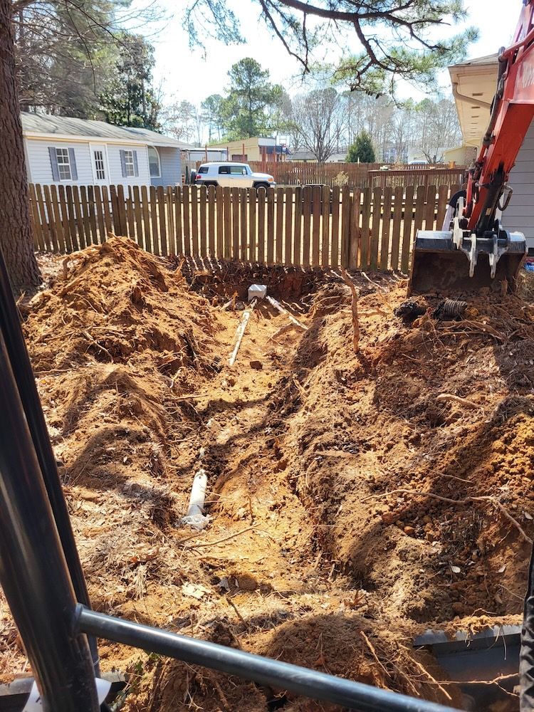 Excavation site with a dug-out dirt pit, piles of soil, a wooden fence, and a backhoe arm on the right.