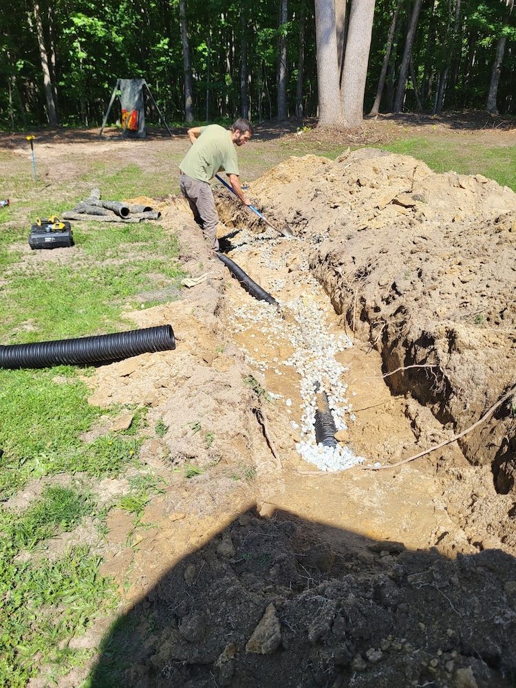 Worker digging a trench beside a yard, with exposed dirt and black drainage pipe along the edge