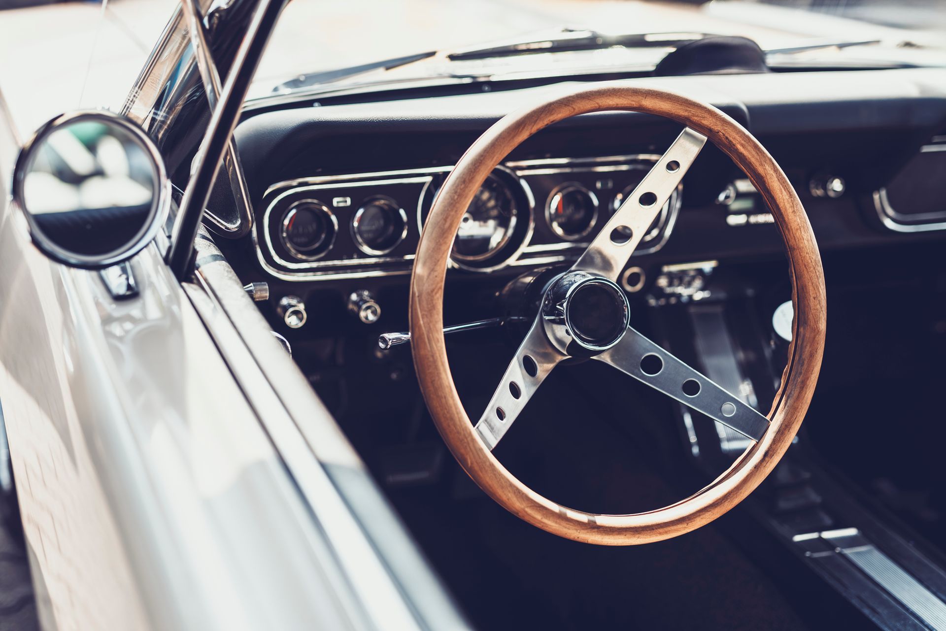 Interior of a classic car: wood steering wheel, chrome accents, instrument panel, and side mirror.