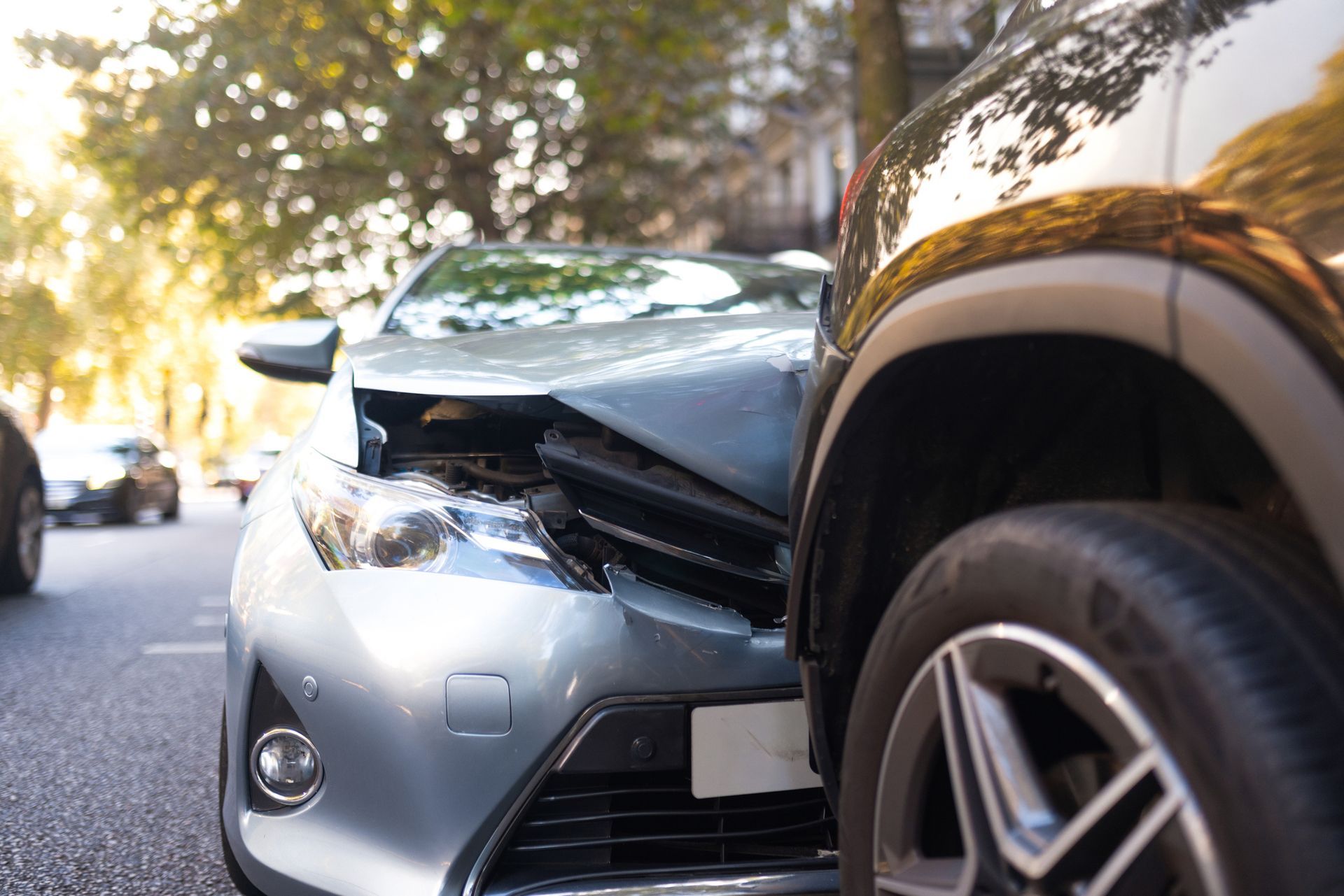 Two cars damaged in a collision on a street; silver car's front crushed, black car's side dented.