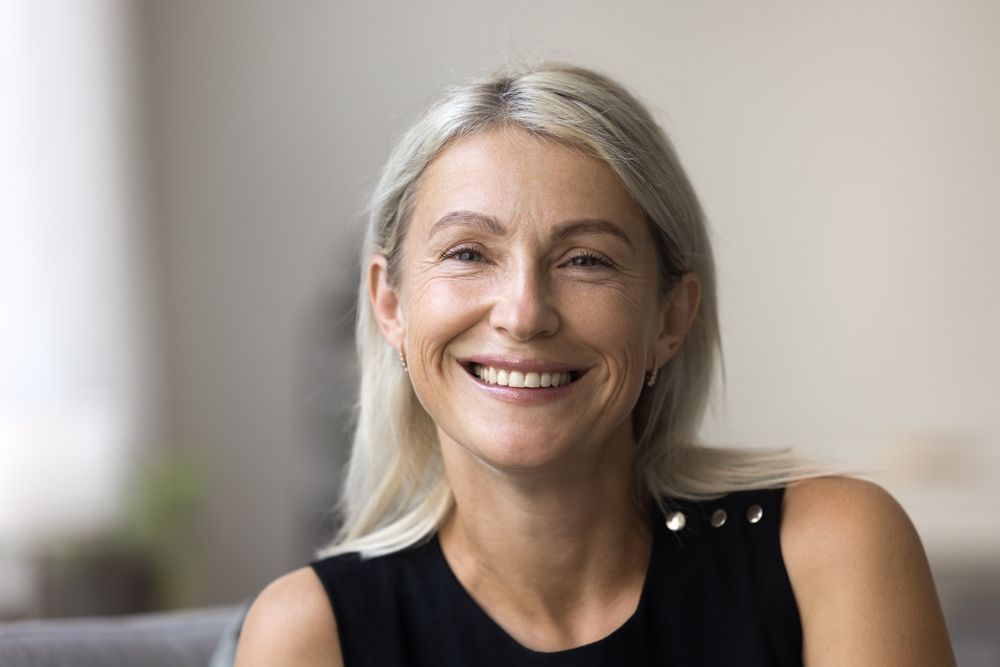 Smiling woman with silver hair in a black sleeveless top, seated indoors against a soft blurred background
