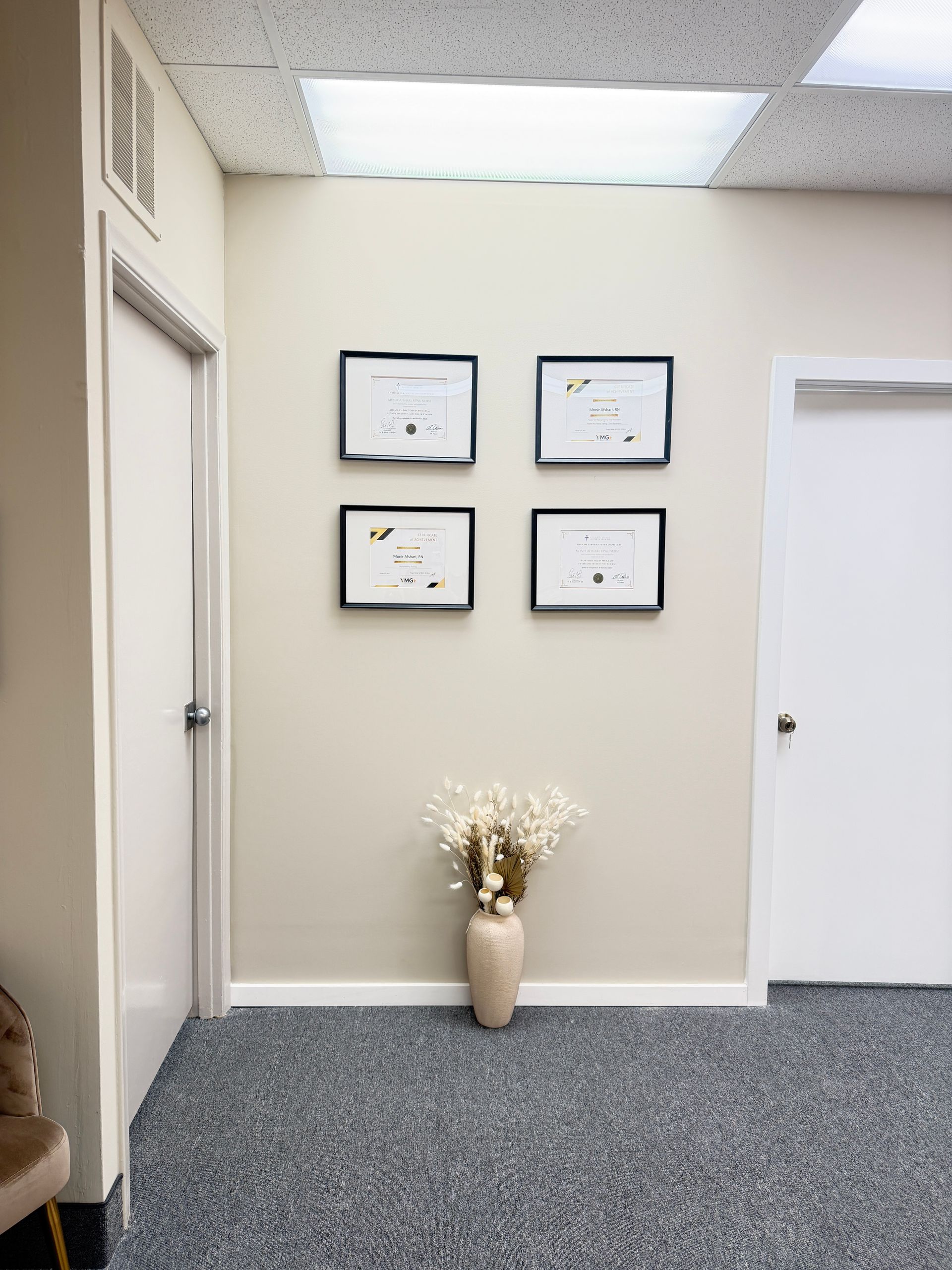 Hallway with framed certificates on a wall above a vase of flowers, carpeted floor, and office doors.