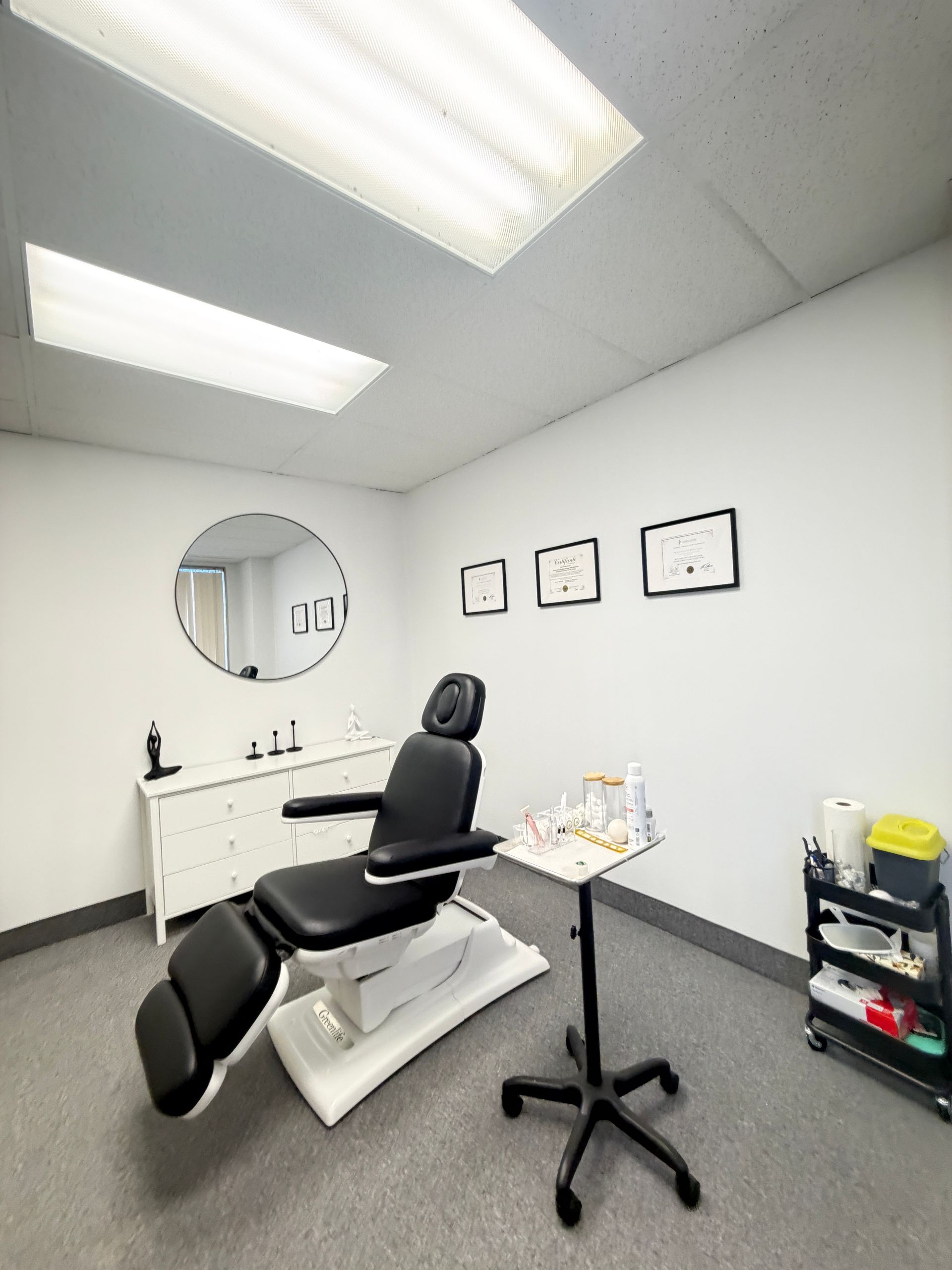 Medical exam room with exam chair, sink, wall art, and a rolling stool under bright overhead lights