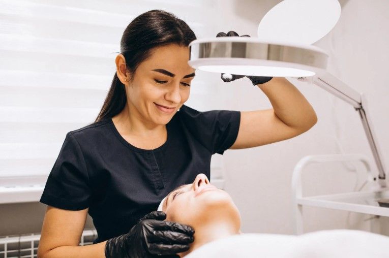 A cosmetologist examines a client’s face under a magnifying lamp in a bright treatment room