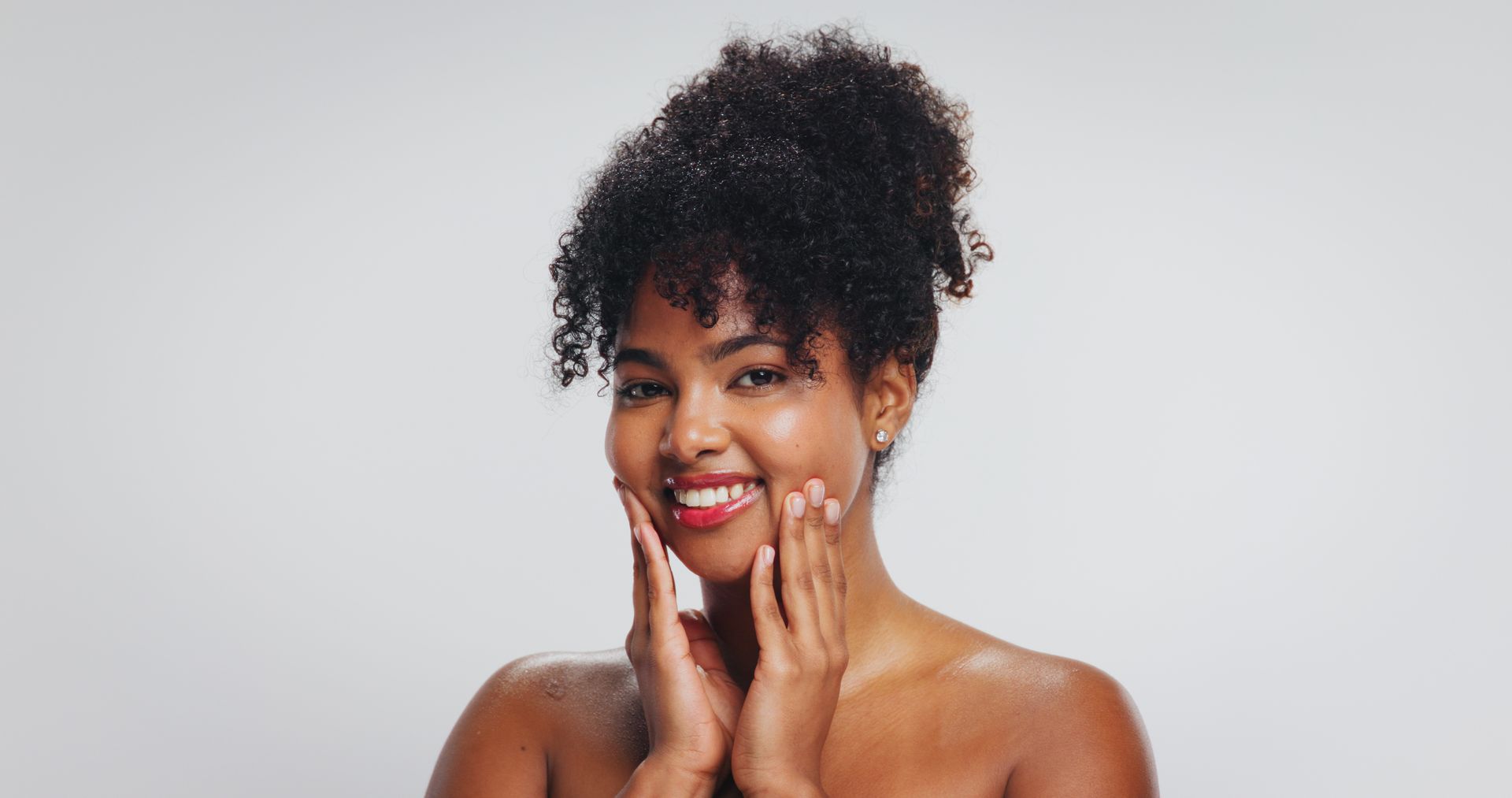 Smiling person with curly hair touching their cheeks against a light gray background