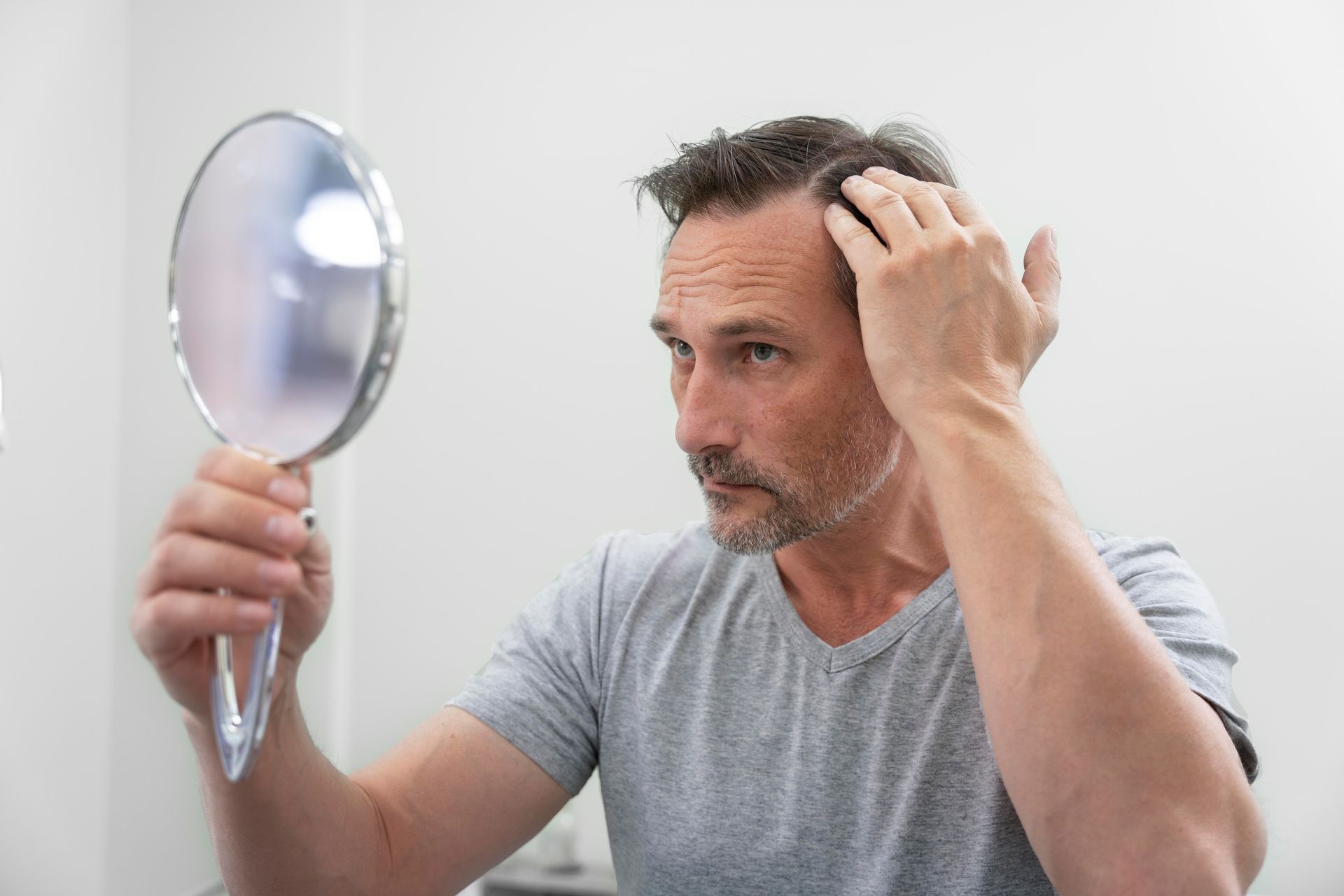 Man examining thinning hair in a mirror, touching his head with a concerned expression.