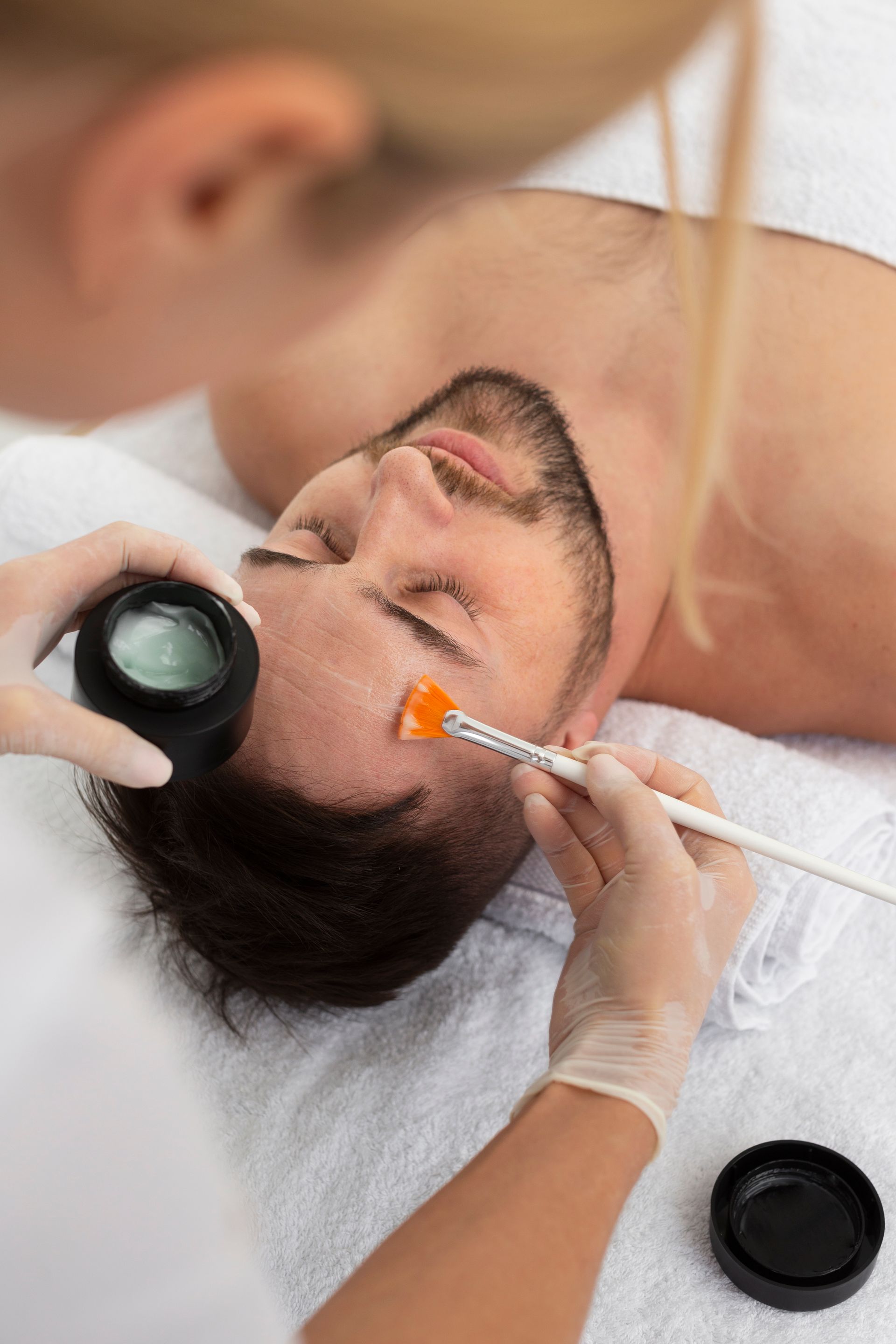 Bearded man receiving facial treatment while lying on a spa bed; esthetician applies orange mask with a brush
