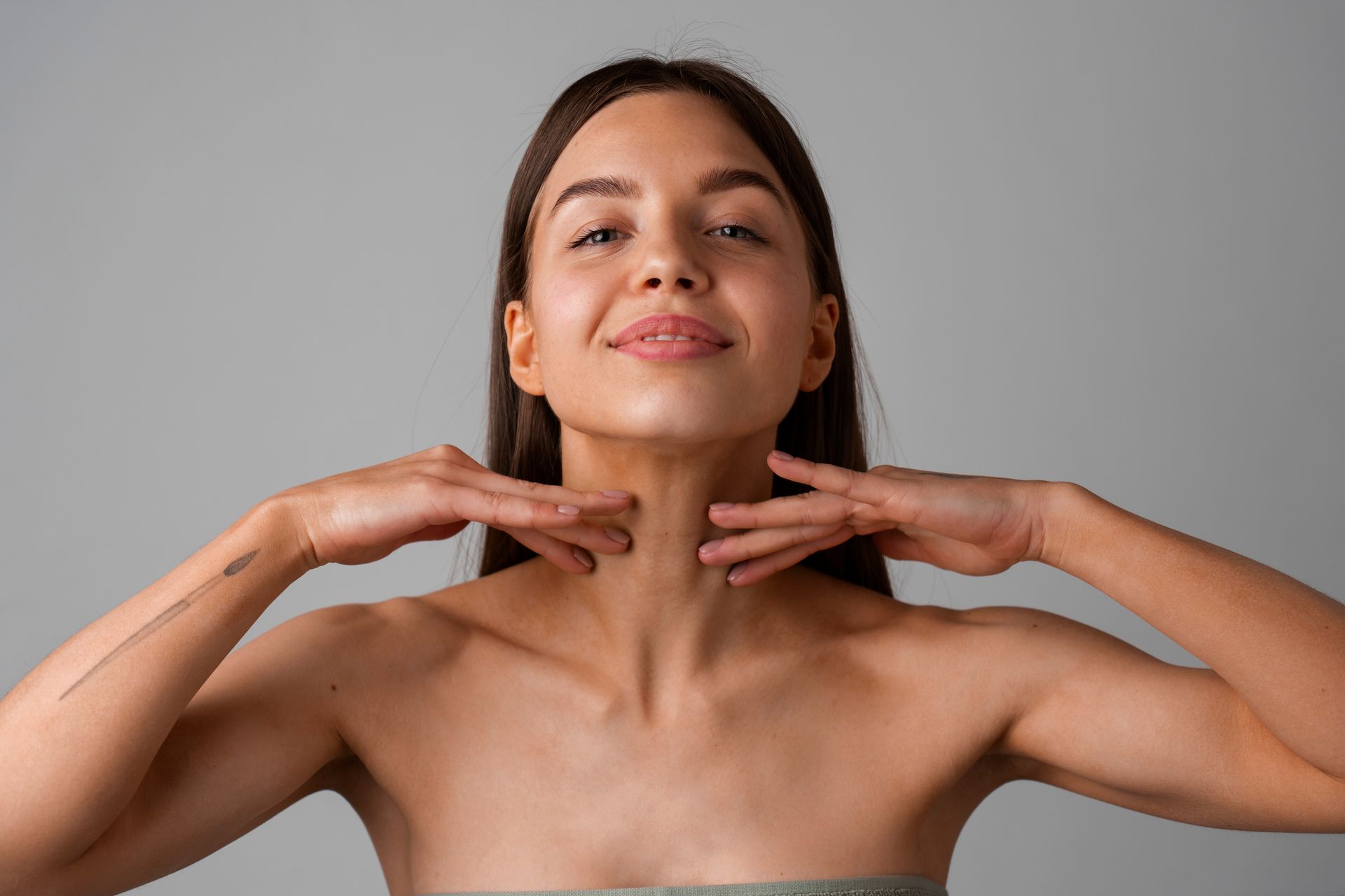 Woman with bare shoulders posing against a gray background, hands framing her neck and a slight smile