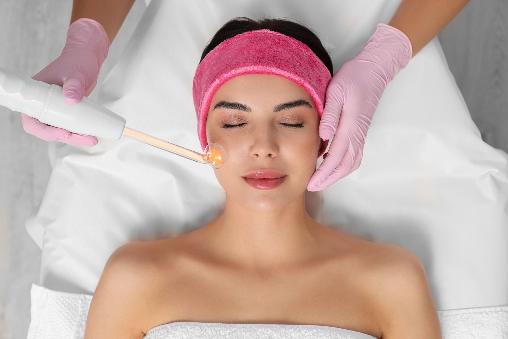 Woman receiving a facial treatment with a pink headband and gloves in a spa setting