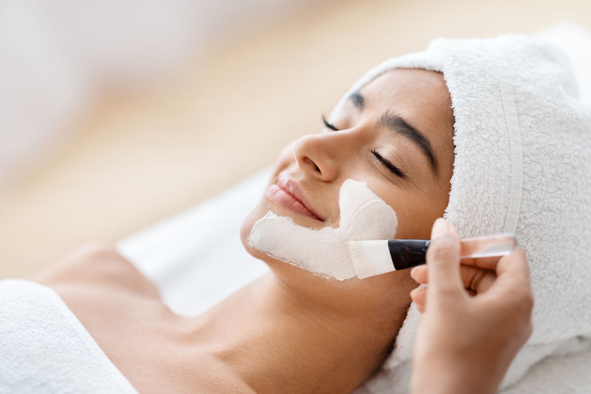 Person receiving a white facial mask treatment with a brush while lying on a spa bed