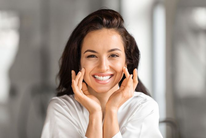 Smiling person in a white shirt resting their face in their hands against a blurred indoor background