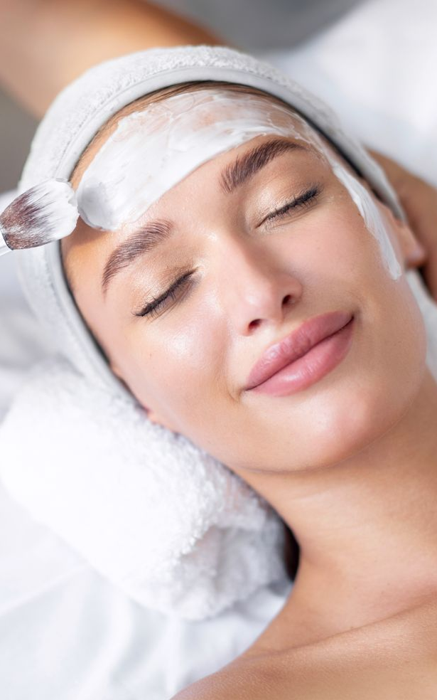 Woman receiving a white facial mask while resting with eyes closed on a towel-covered bed