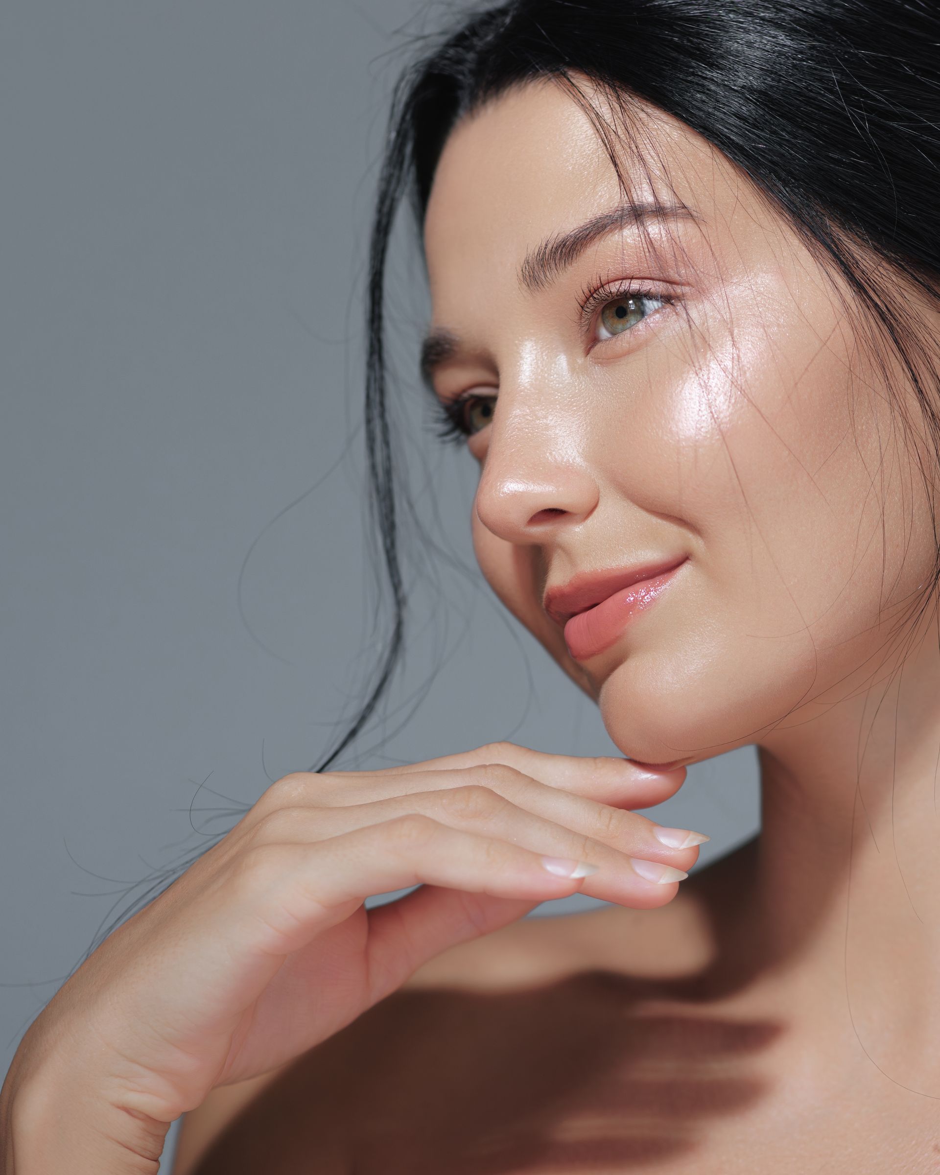 Close-up portrait of a smiling woman with dewy skin, resting her hand under her chin against a gray background