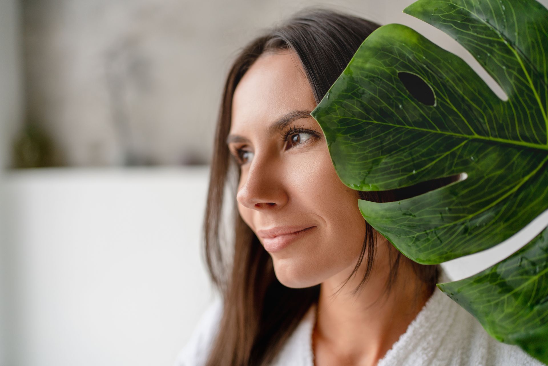 Woman in a white sweater looking sideways beside a large green leaf indoors