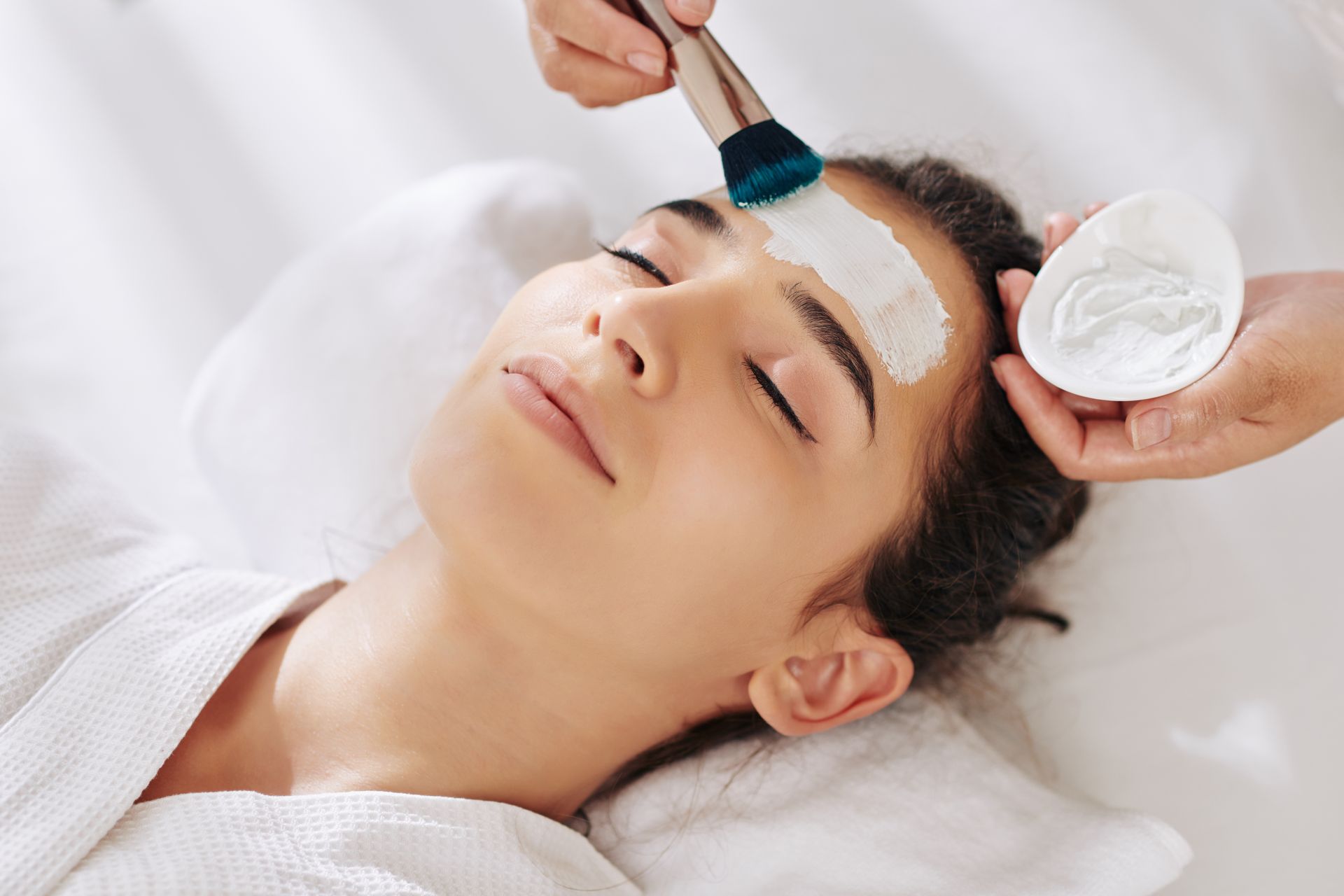 Spa treatment applying white facial mask with brush while a woman relaxes with eyes closed