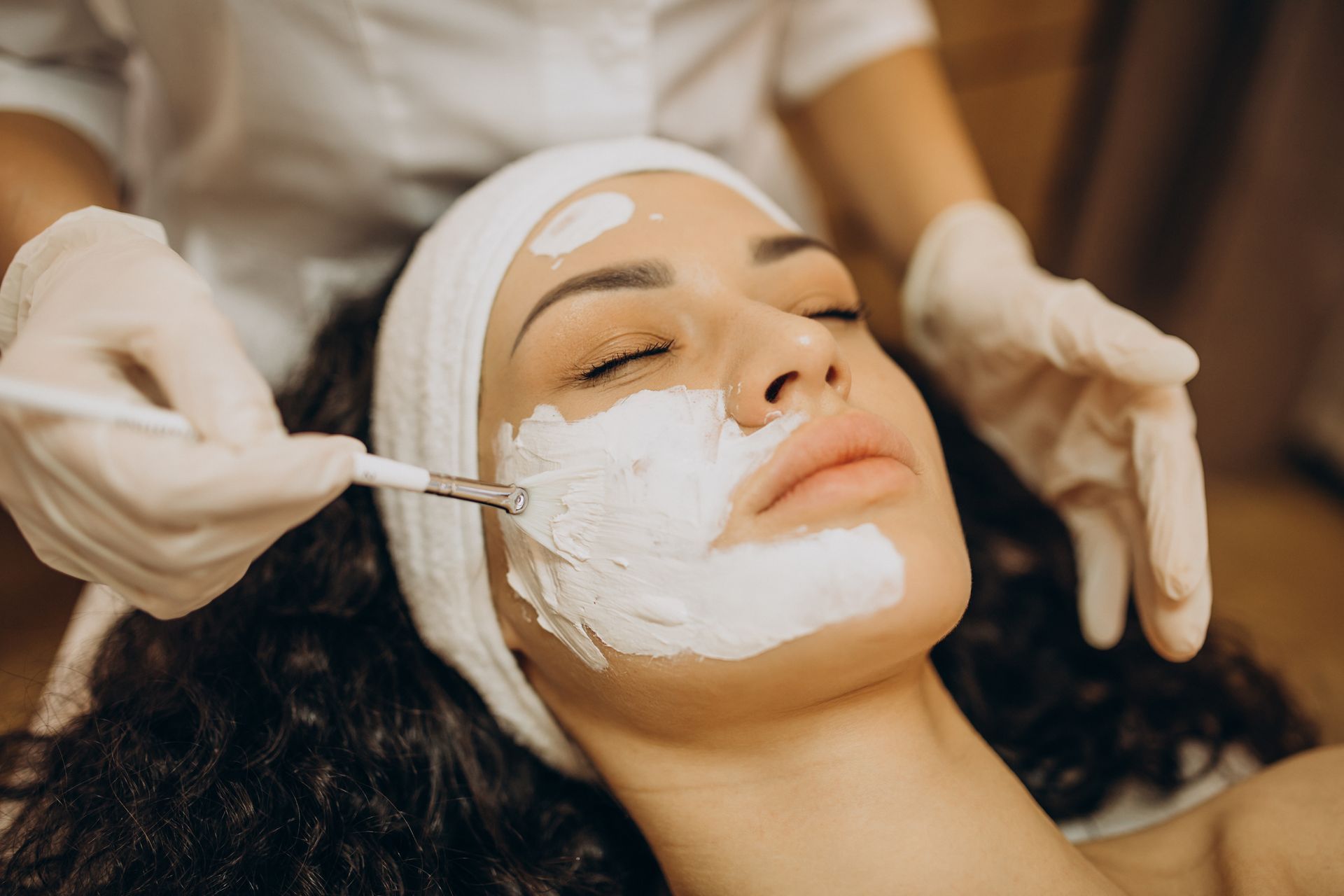 Spa esthetician applying a white facial mask to a woman lying back with eyes closed