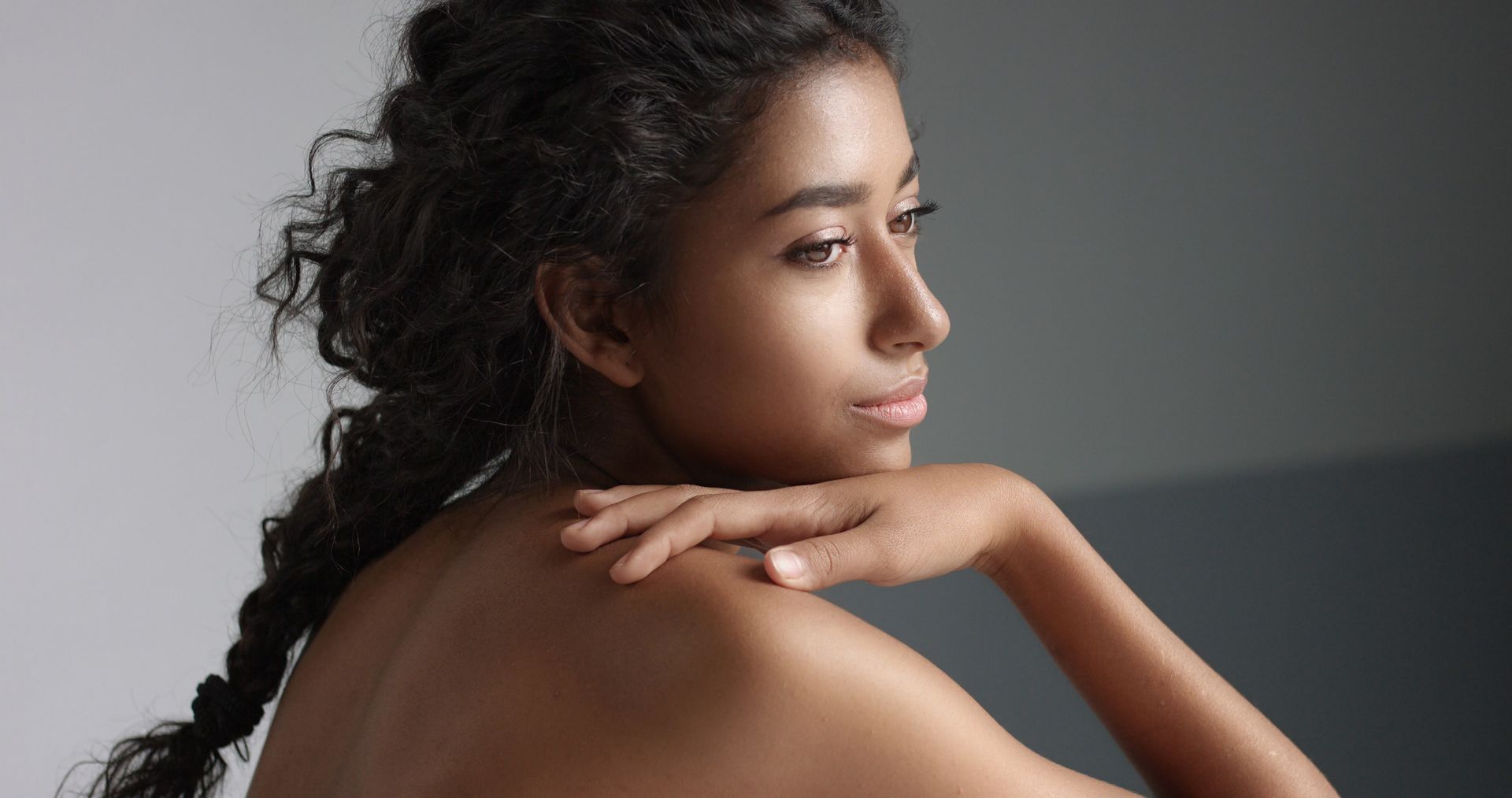 Woman with curly hair resting her chin on her hand, looking over her shoulder against a gray background