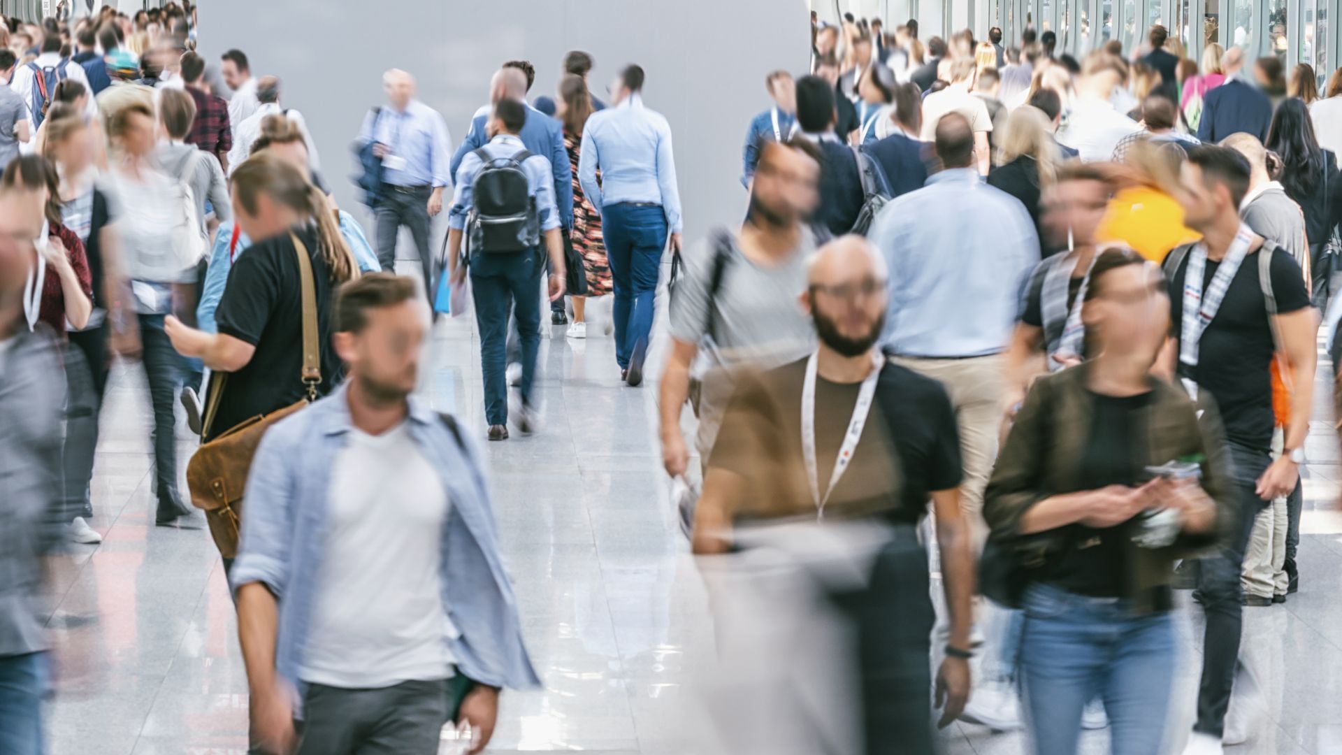 Crowd of people walking in a brightly lit hallway. Many are blurred, some wearing lanyards, casual attire.