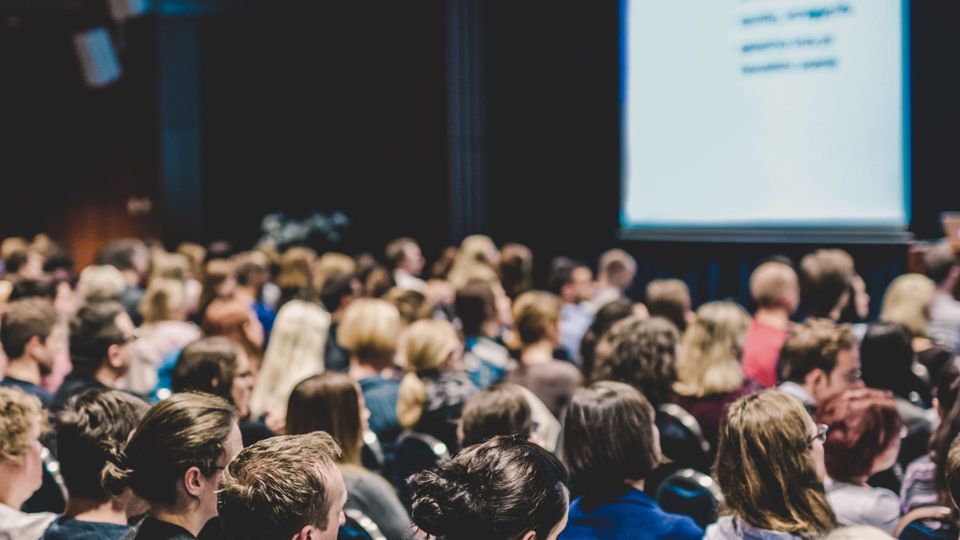Audience seated in a large auditorium, facing a screen displaying text or a presentation.
