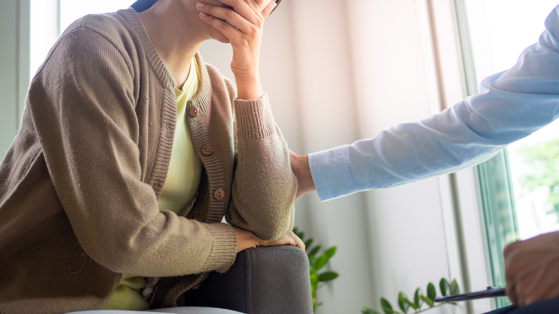 Woman in distress, hand covering her mouth, receiving comfort from a person's hand on her shoulder.