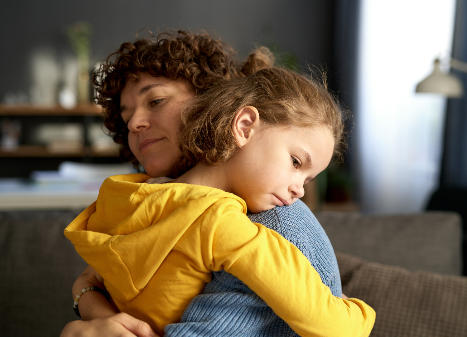 Therapist with clipboard consults a young girl on a couch, holding a teddy bear. Bright room.