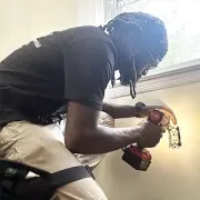 Person drilling a wall near a window, wearing a black shirt and beige shorts, with dust and debris visible