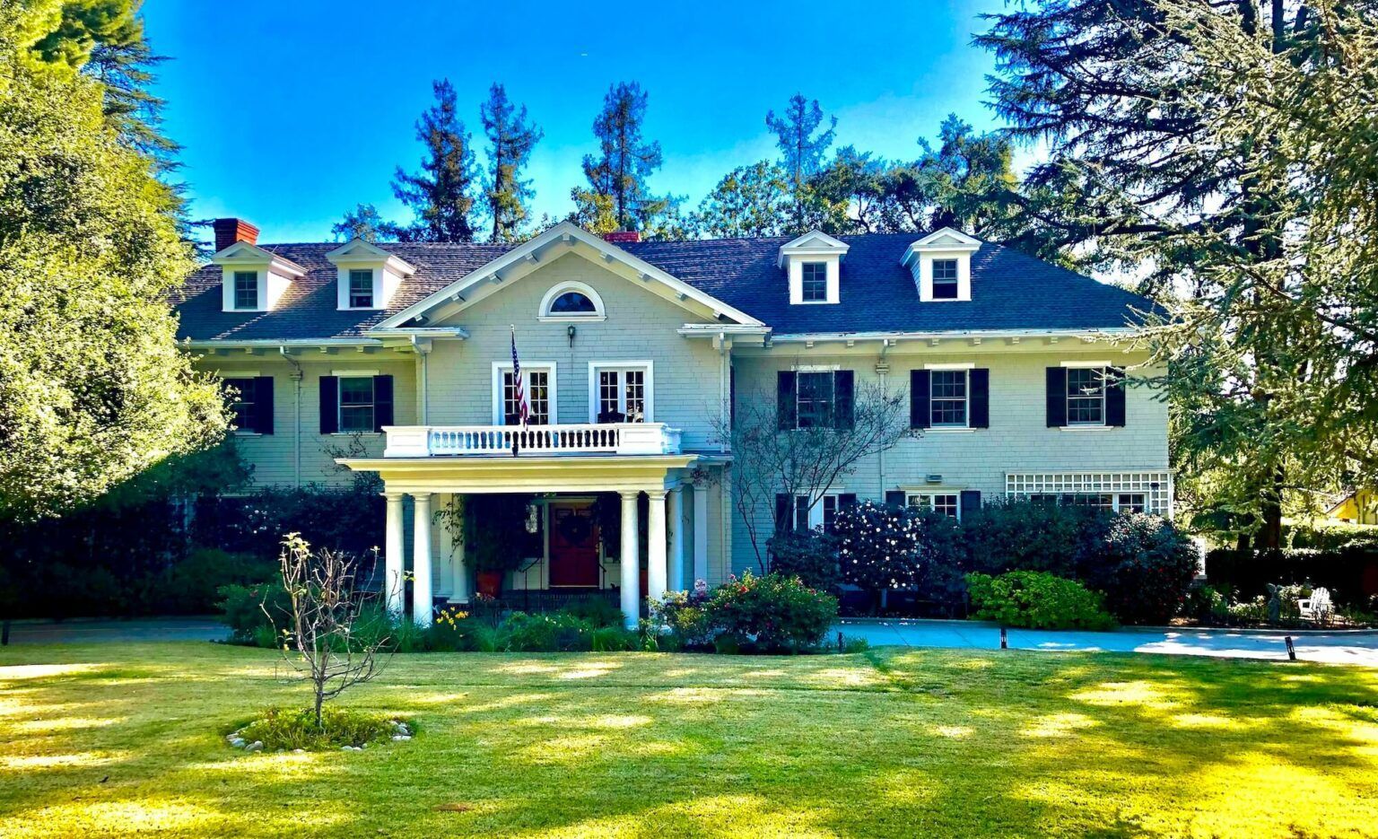 A two-story light gray house with a white porch and dormers, set on a green lawn with trees.