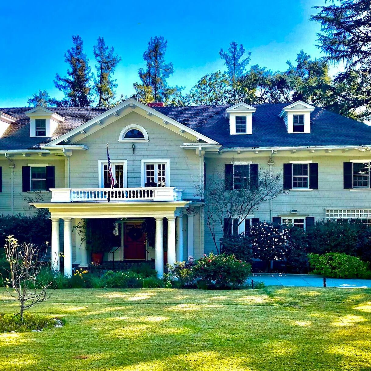 Two-story light gray house with white columns, balcony, black shutters, and green lawn under a blue sky.