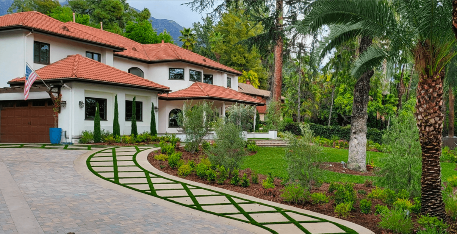 White mansion with red tile roof, surrounded by green landscaping and palm trees. Driveway with stone and grass.