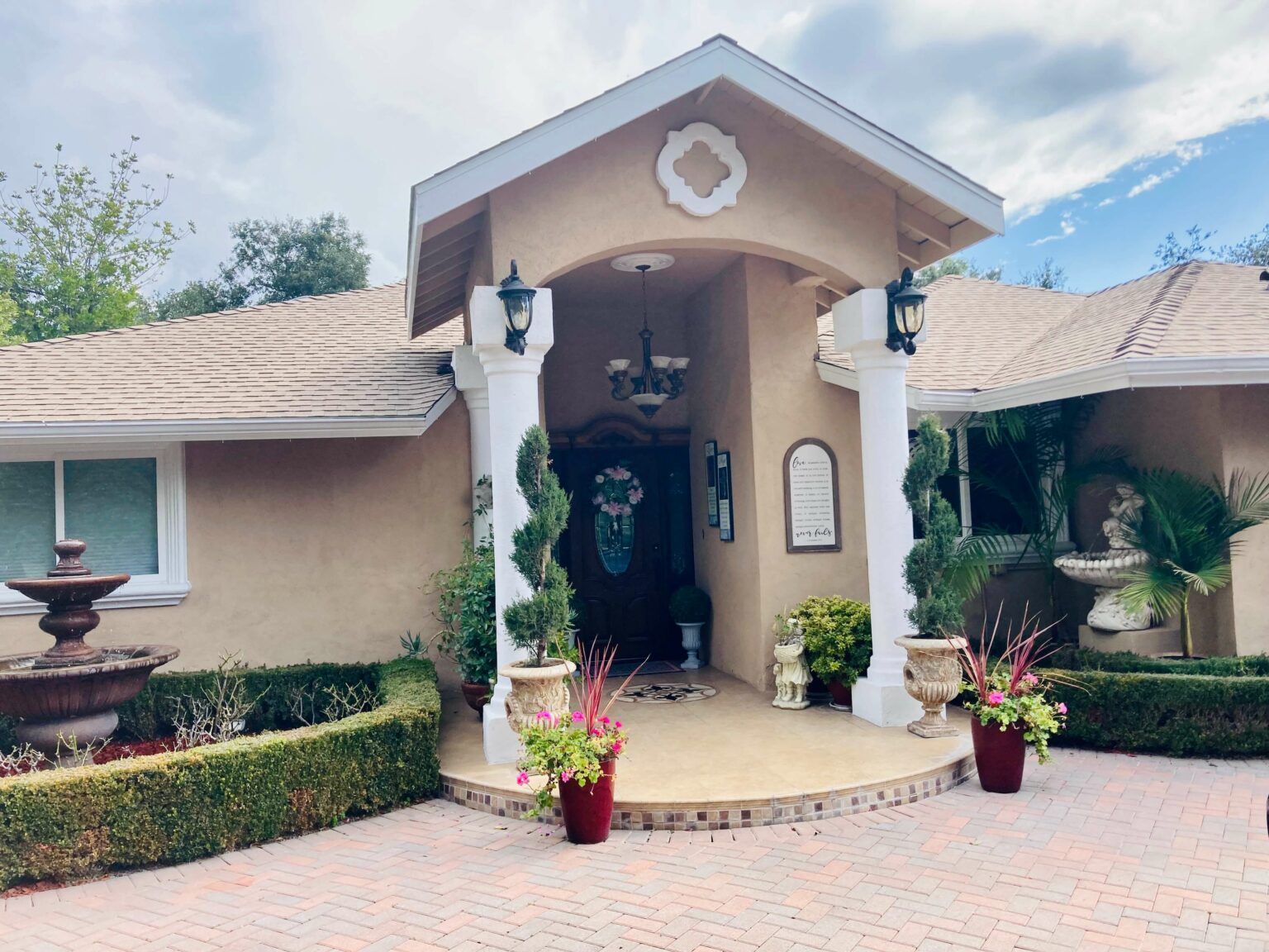Tan house with ornate columns, arched entryway, and red brick driveway.