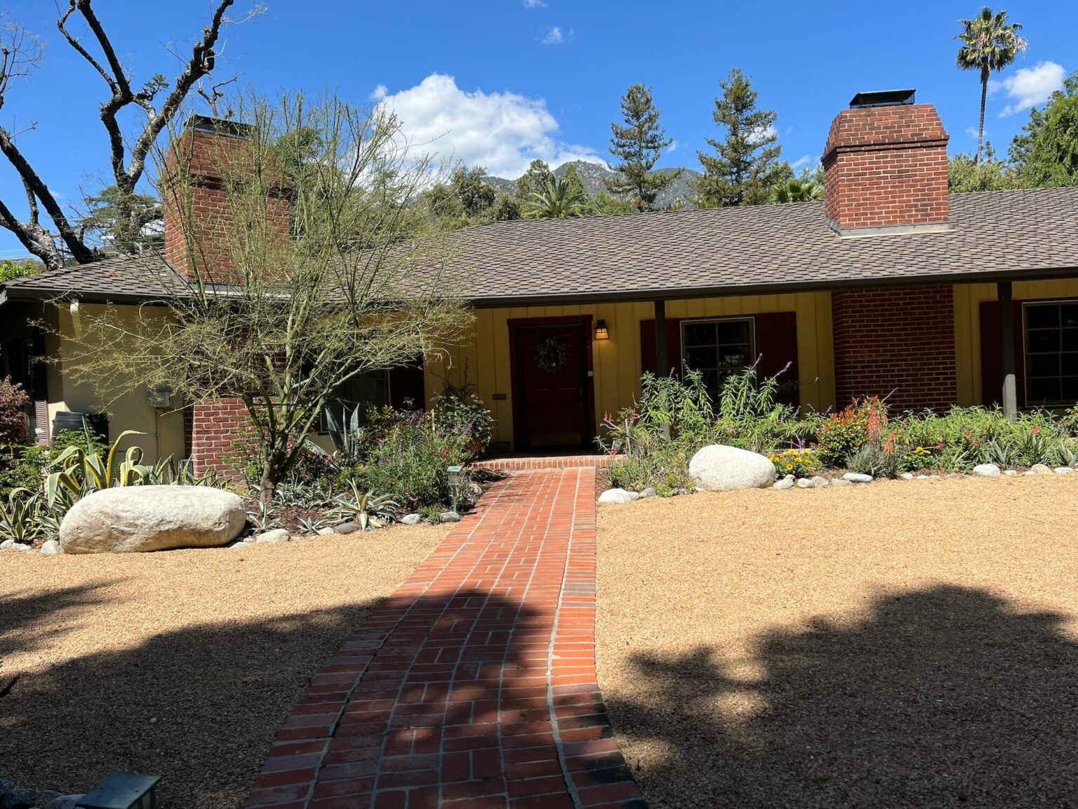 A brick pathway leads to a yellow house with a red door and chimneys; landscaping includes rocks and plants.