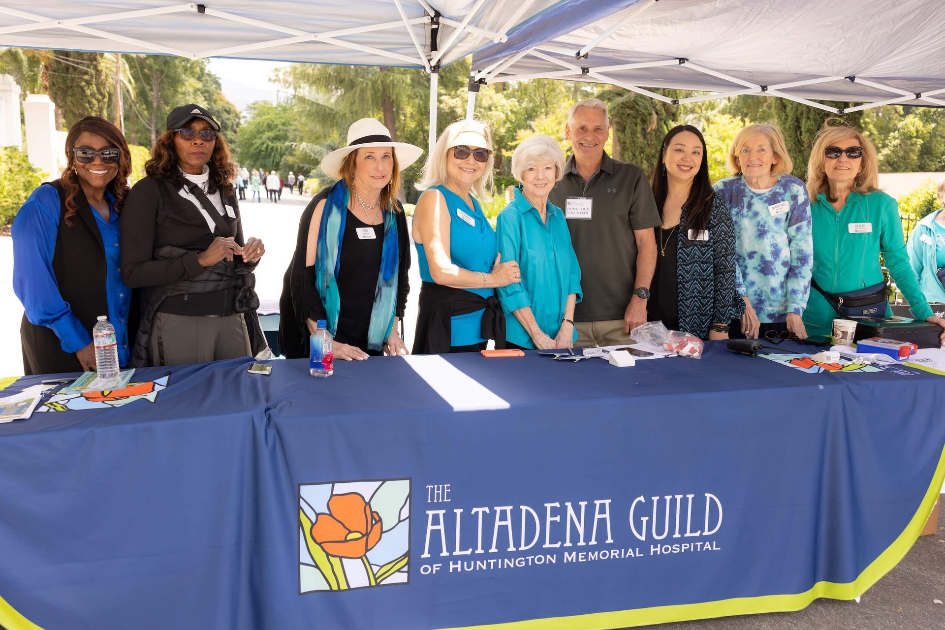 People at a table with "Altadena Guild" logo. They stand behind a blue table, smiling outdoors.