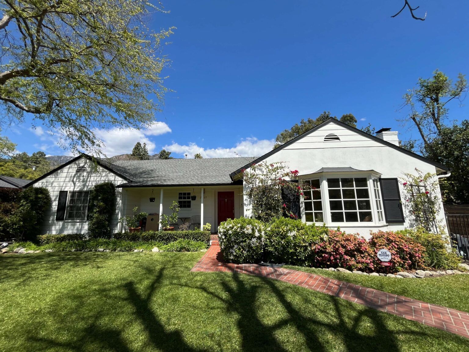 White house with black shutters, red door, brick walkway, and green lawn under a blue sky.